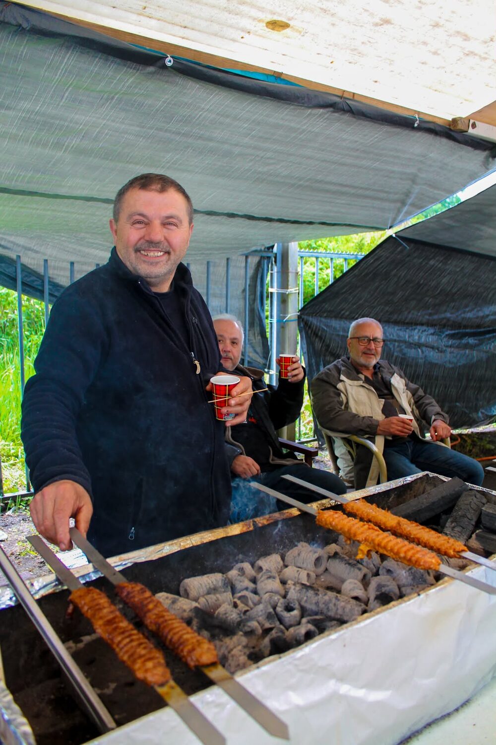 Een lachende vrijwilliger draait de kebabspiesen op de houtskoolgrill